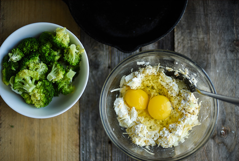 Chopped broccoli in small bowl next to larger bowl with cheese and raw eggs.