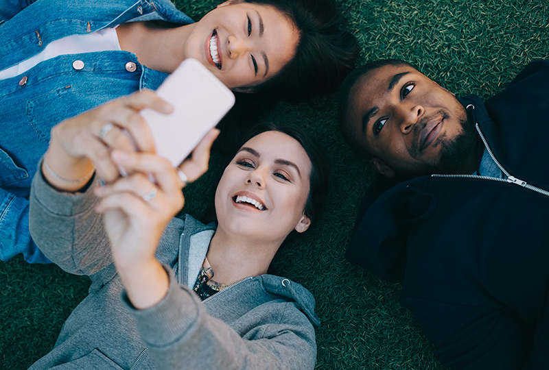 Three teens lying on the grass taking selfies on a cellphone