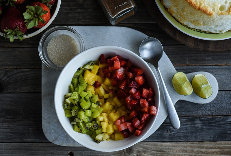 Chopped strawberries, pineapple and kiwi in a mixing bowl, with a small bowl of sugar nearby.