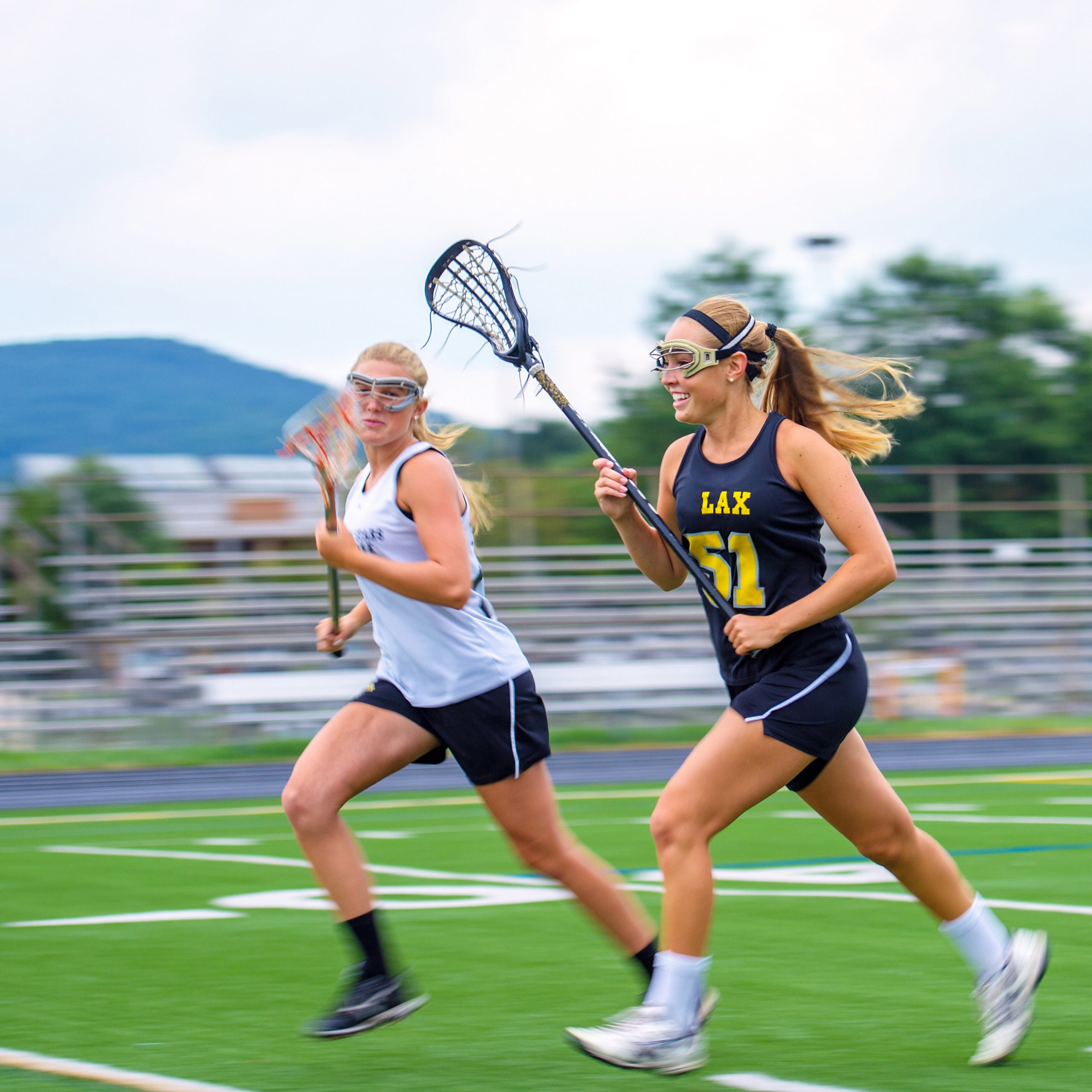Two teenaged girls playing lacrosse on an outdoor field wearing eye protection.