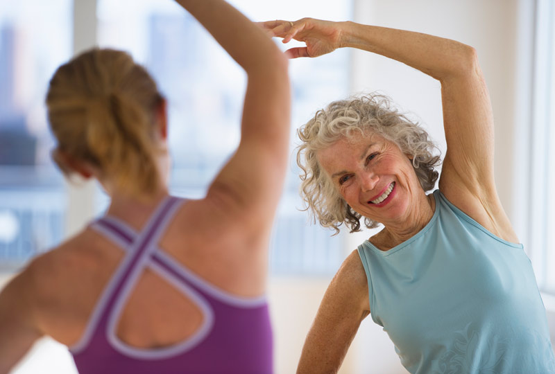 Older woman follows a younger routine by a younger instructor in stretching exercise.