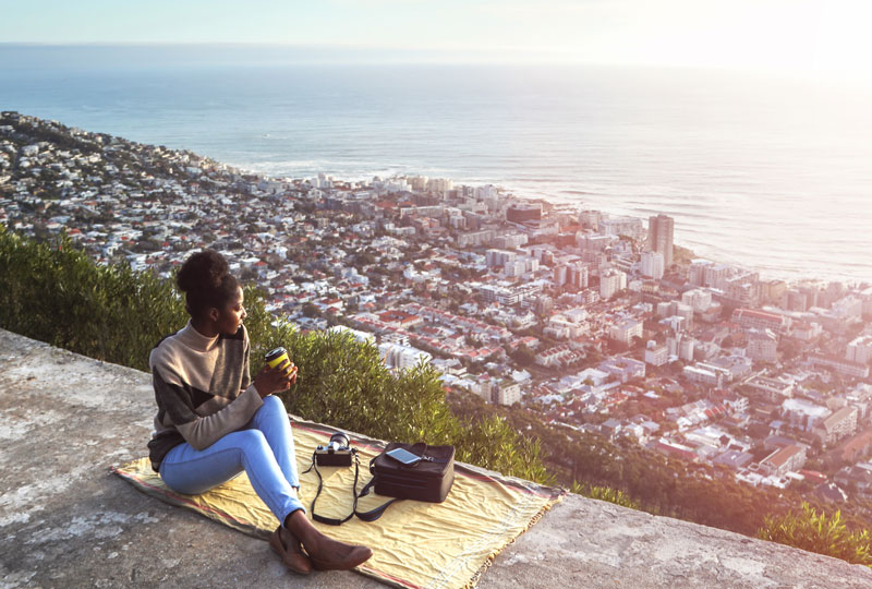 A woman sits on an overlook with the view below of a large city she has traveled to.