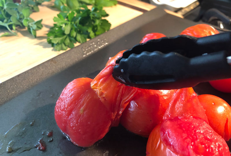 Lightly roasted whole tomatoes on a baking sheet; tongs are peeling them
