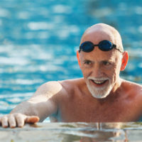 A smiling older man stands in chest-deep water at the edge of a swimming pool.