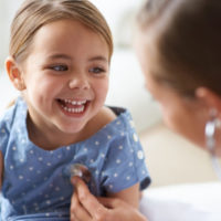 A young girl smiles at a healthcare provider, who is listening to her heartbeat.