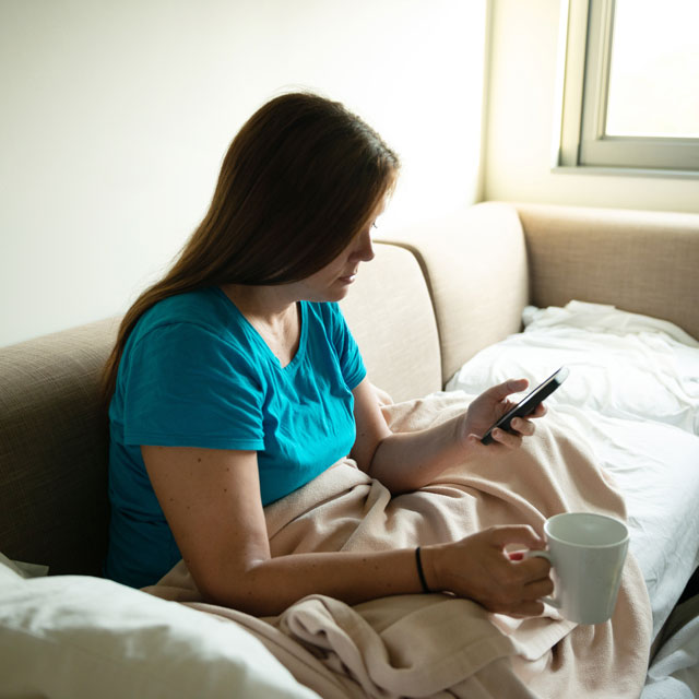 Woman sits on couch with blankets, holding cell phone and a mug of coffee.
