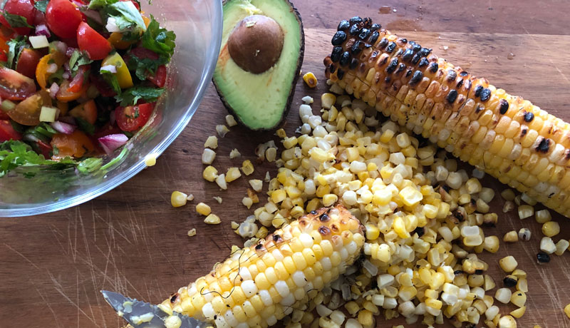 Grilled corn on a cutting board with half an avocado and a bowl of salsa being mixed.