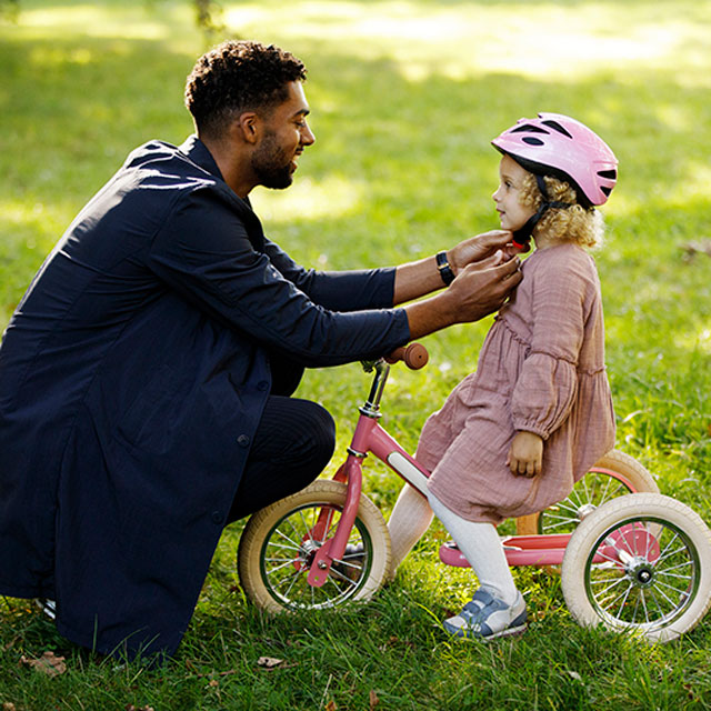 A father helps his young daughter put on a bicycle helmet as she sits on a tricycle.