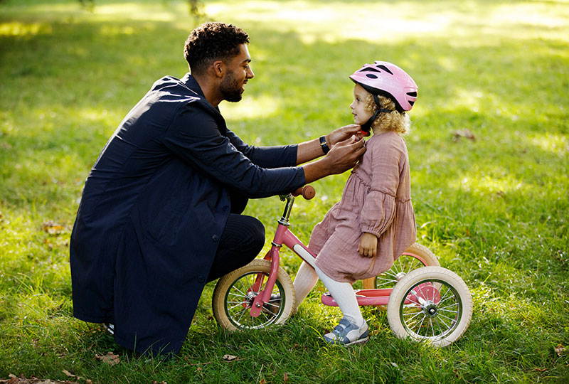 A father helps his young daughter put on a bicycle helmet as she sits on a tricycle.