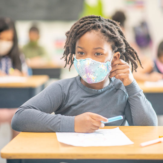 6-year-old girl wearing a mask sits at a school desk in a classroom.