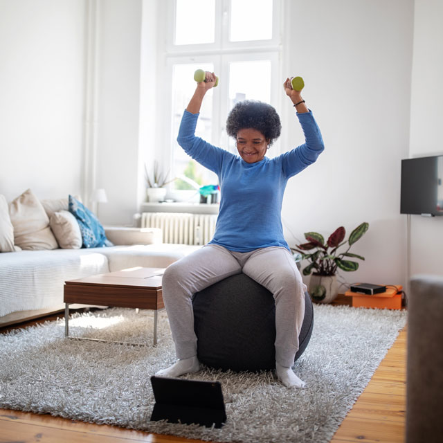 Woman does overhead shoulder presses holding small dumbbells while sitting on an exercise ball watching an online workout video.