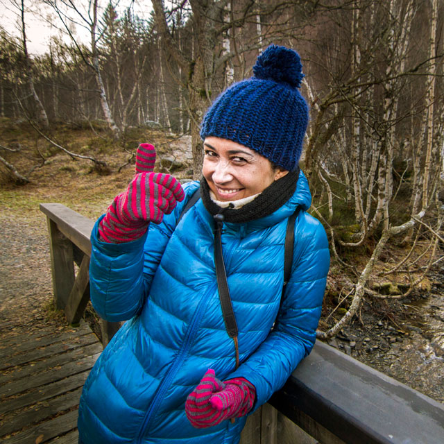 A woman dressed in a parka, hat and gloves stands on a wood bridge over a creek in the woods in wintertime.