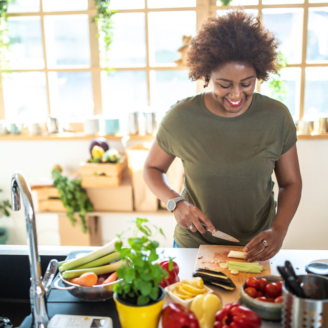 An African-American woman chops fresh vegetables in a sunny kitchen.
