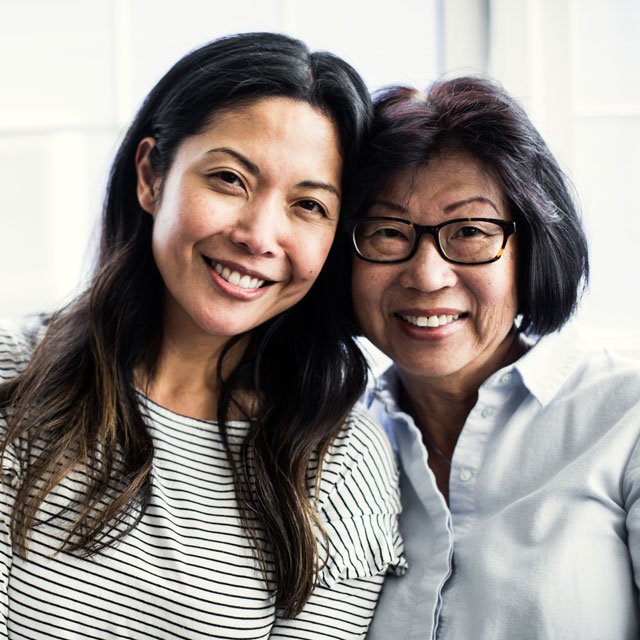 A young Asian woman stands close to her mother as they both look into the camera.