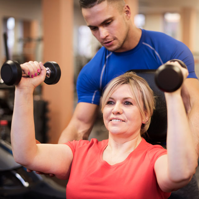 A woman is seated in a gym doing shoulder presses with two dumbbells with a male trainer helping her.