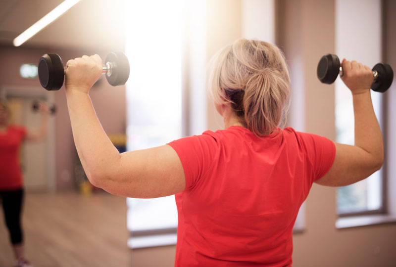 A woman does shoulder presses with both arms simultaneously holding two dumbbells while watching in the mirror.