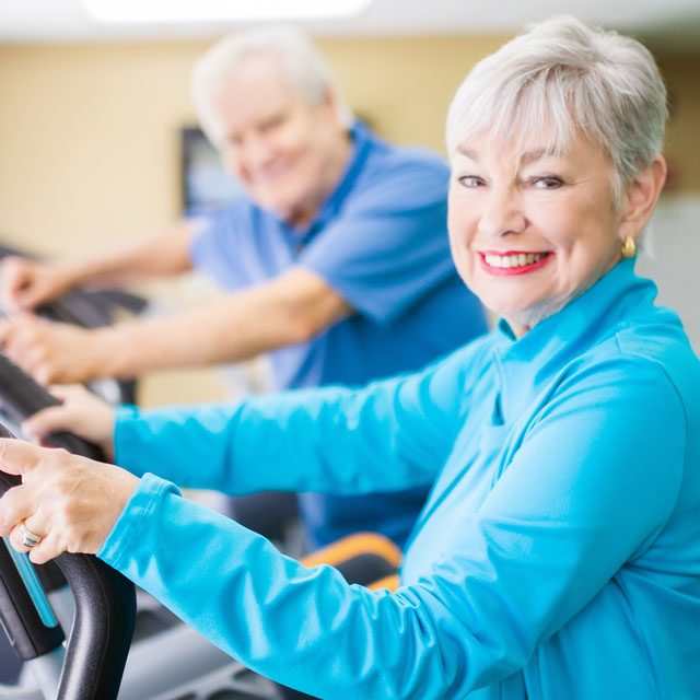 An older couple use elliptical machines in a gym.
