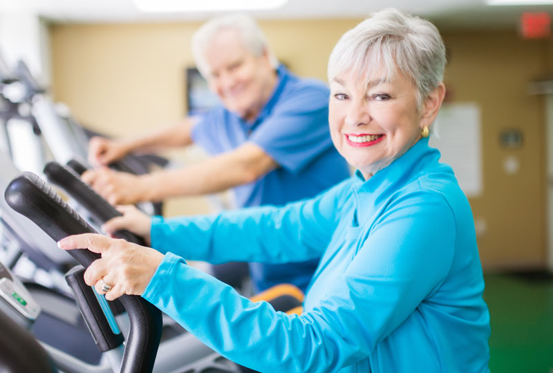An older couple use elliptical machines in a gym.