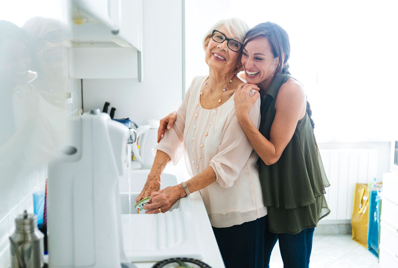 A younger woman hugs an older woman who is washing dishes in a kitchen sink.