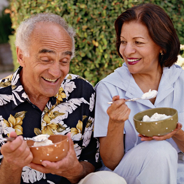 An older Latino couple sit outdoors, eating ice cream from large bowls.