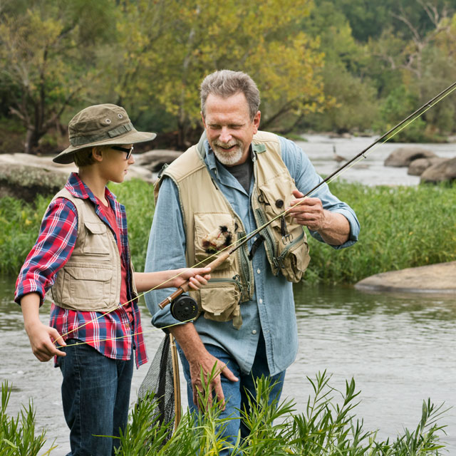 A middle aged man teaches a boy how to fly fish, standing on the banks of a river.