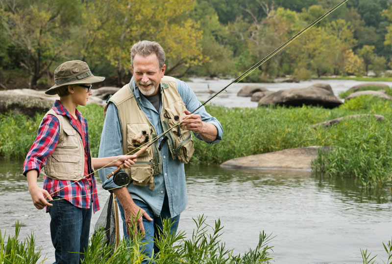 A middle aged man teaches a boy how to fly fish, standing on the banks of a river.