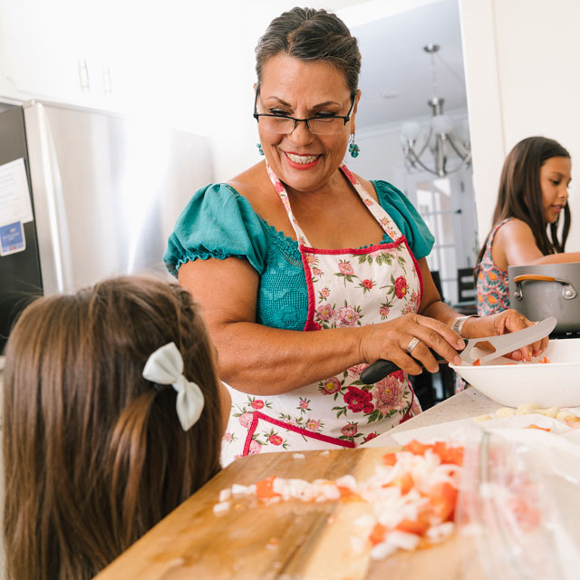 A middle aged Latina woman prepares food in the kitchen with two girls helping.