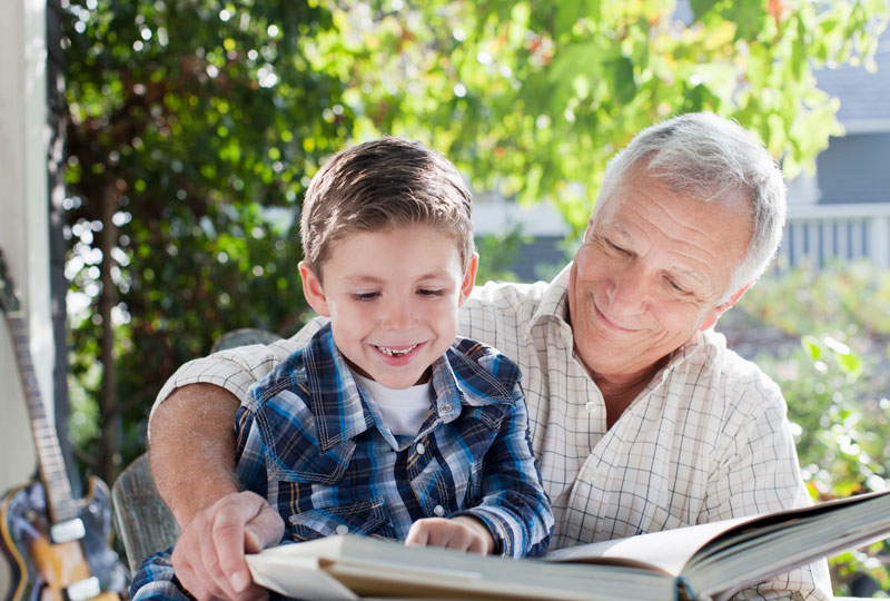 An older man reads a book with a boy as they are sitting outdoors.