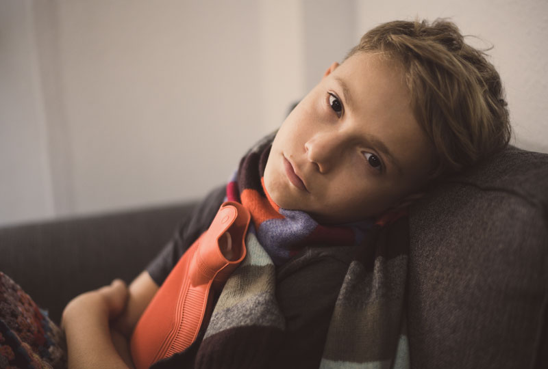 A school-aged boy sits on a couch with a hot water bottle pressed to his stomach
