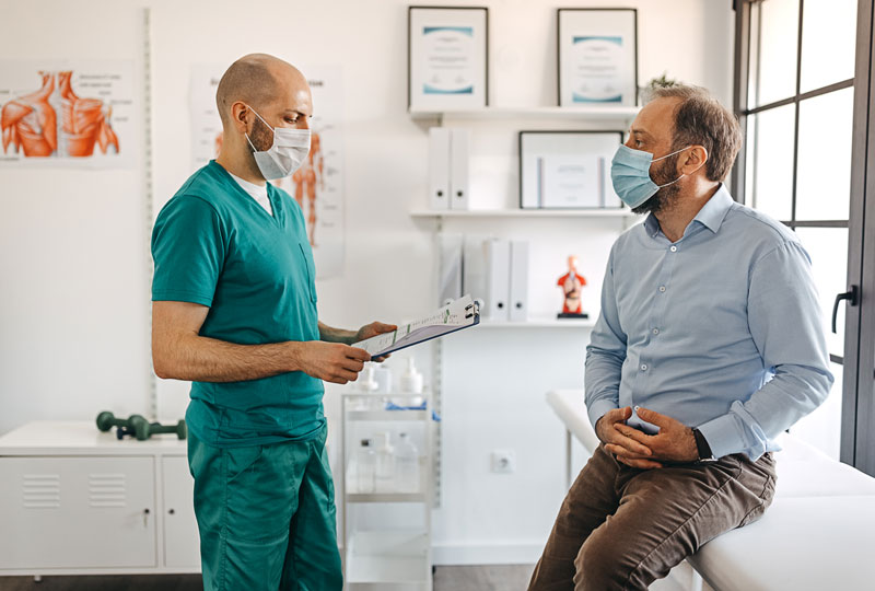 A male doctor in scrubs consults with a male patient in a clinic room, both wearing masks.