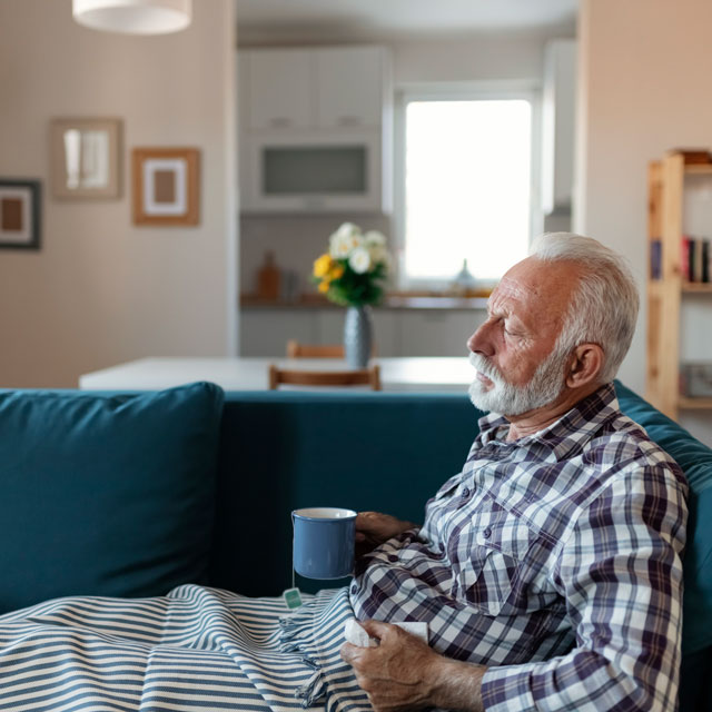 An older man sits on a couch under a blanket with a mug in one hand, looking fatigued.