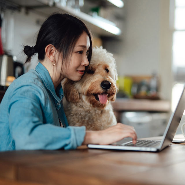 Woman cuddling dog and working at laptop in kitchen, practicing work-life balance strategies