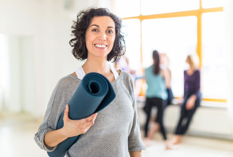 Woman stands in an exercise studio holding a yoga mat.