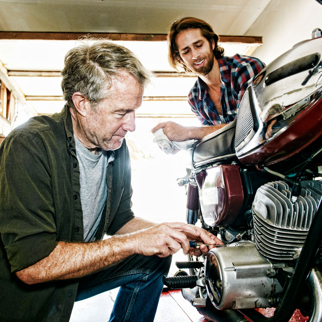 An older man kneels to work on a motorcycle engine while a younger man watches in a garage.