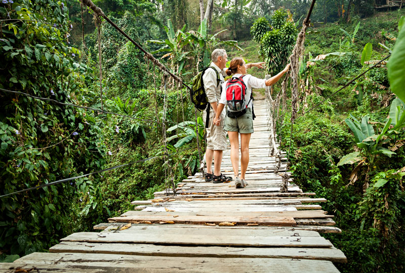 White middle aged couple stand on a plank bridge in a jungle.