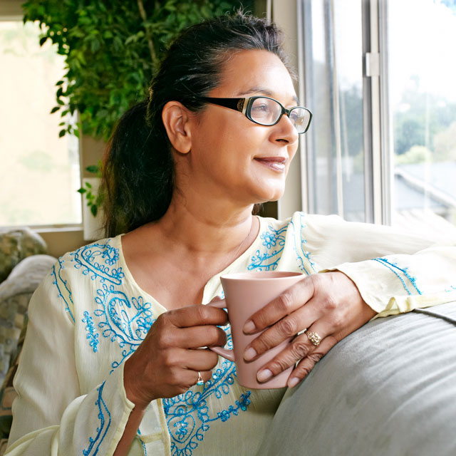 Head-and-shoulders photo of a mixed-race woman holding a mug of coffee while sitting by a window.