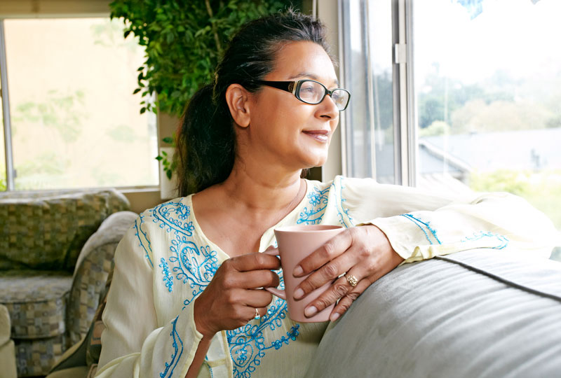 Head-and-shoulders photo of a mixed-race woman holding a mug of coffee while sitting by a window.
